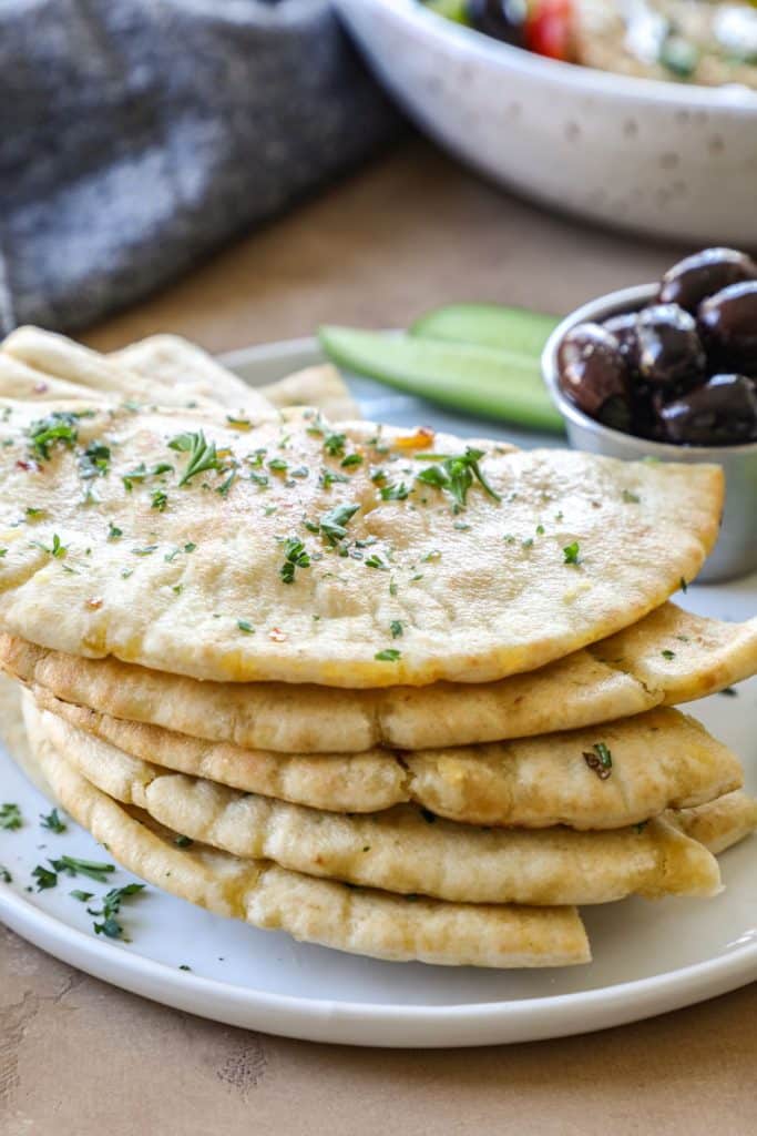 Pita bread stacked on a white plate with a bowl of olives in the background.