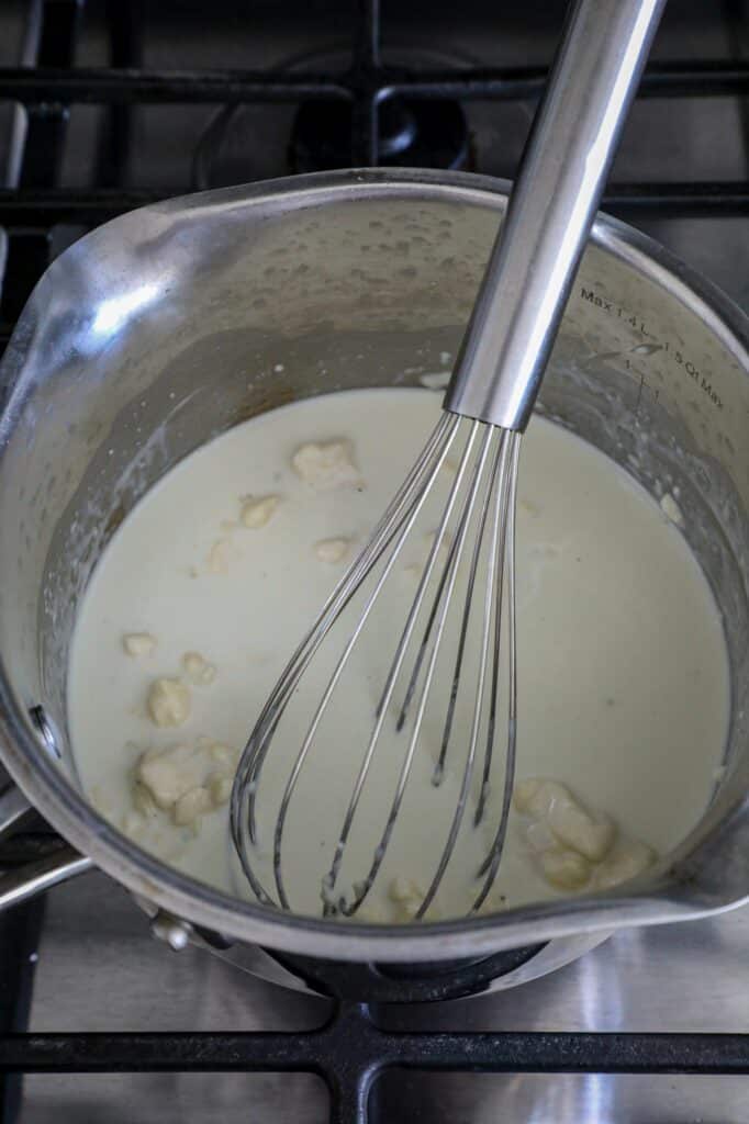 Cream and blue cheese in a silver sauce pot with a whisk in it.